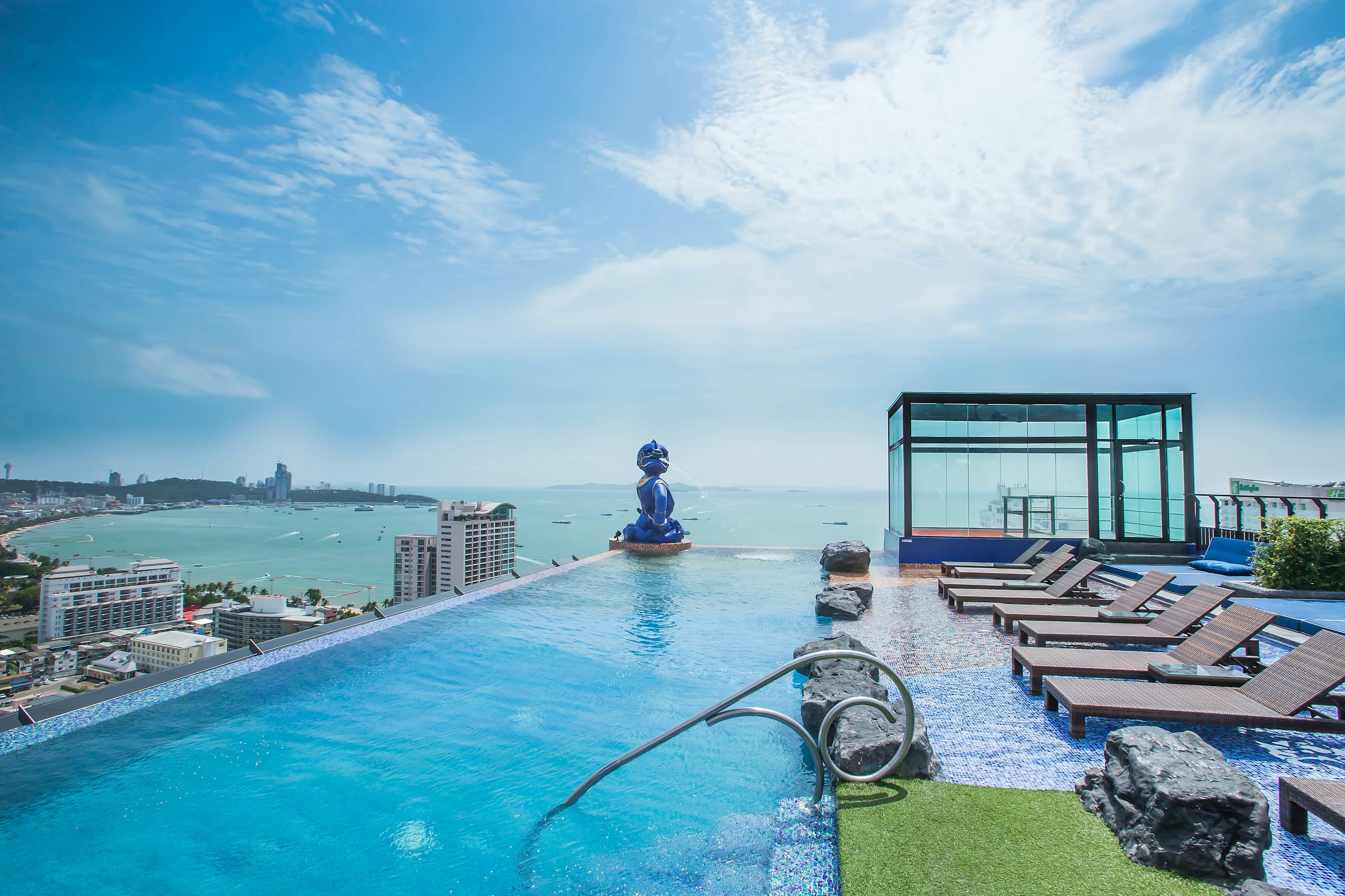 Rooftop infinity pool with a blue statue overlooking a coastal city and ocean bay.